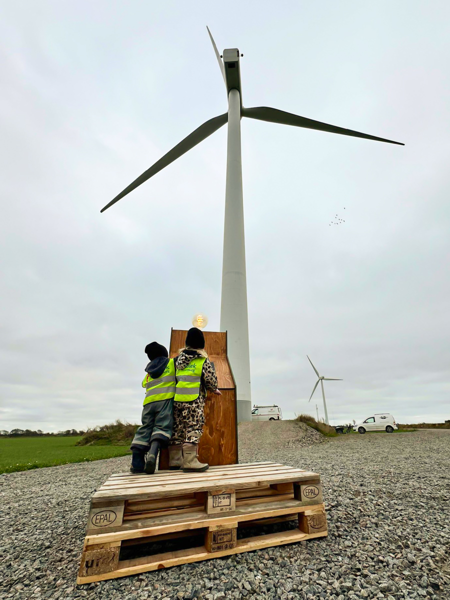 Två små barn i varselvästar står på en träpall med ryggen mot kameran och drar i en invigningsspak framför ett vindkraftverk.