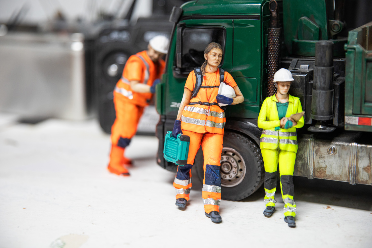 Tre små dockor med säkerhetskläder står framför en leksakslastbil