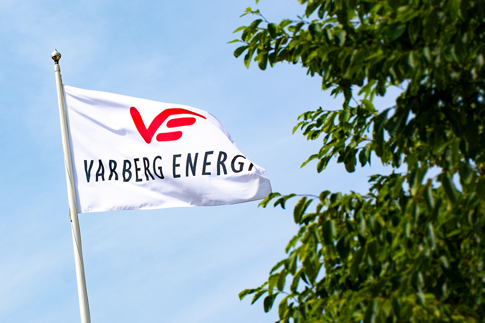 Vit flagga med Varberg Energis logotype på med blå himmel bakom och ett grönt träd bredvid