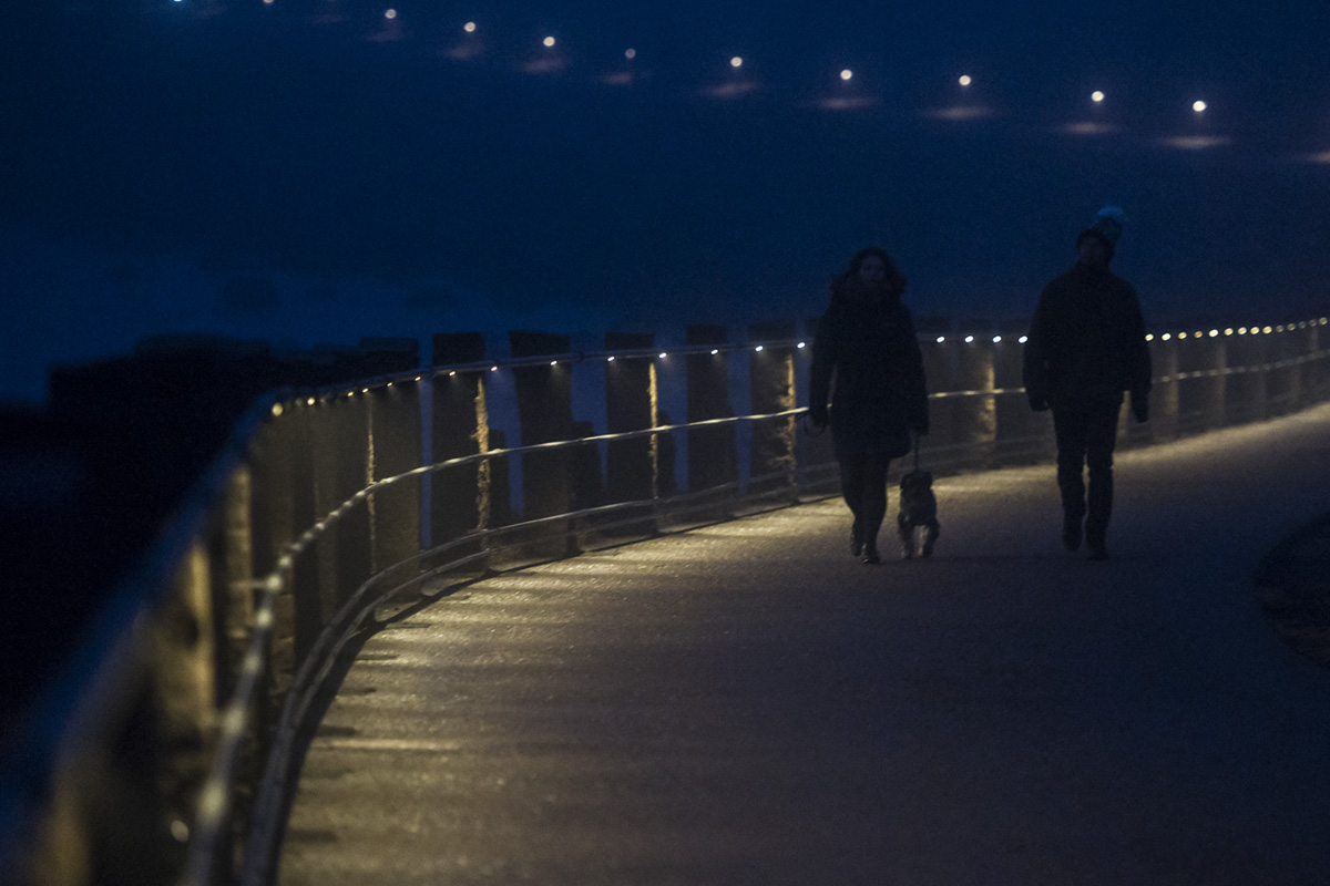 Två personer med hund går i mörkret på strandpromanden i mörker och ljuskällorna i staketet mot havet lyser upp gångvägen.