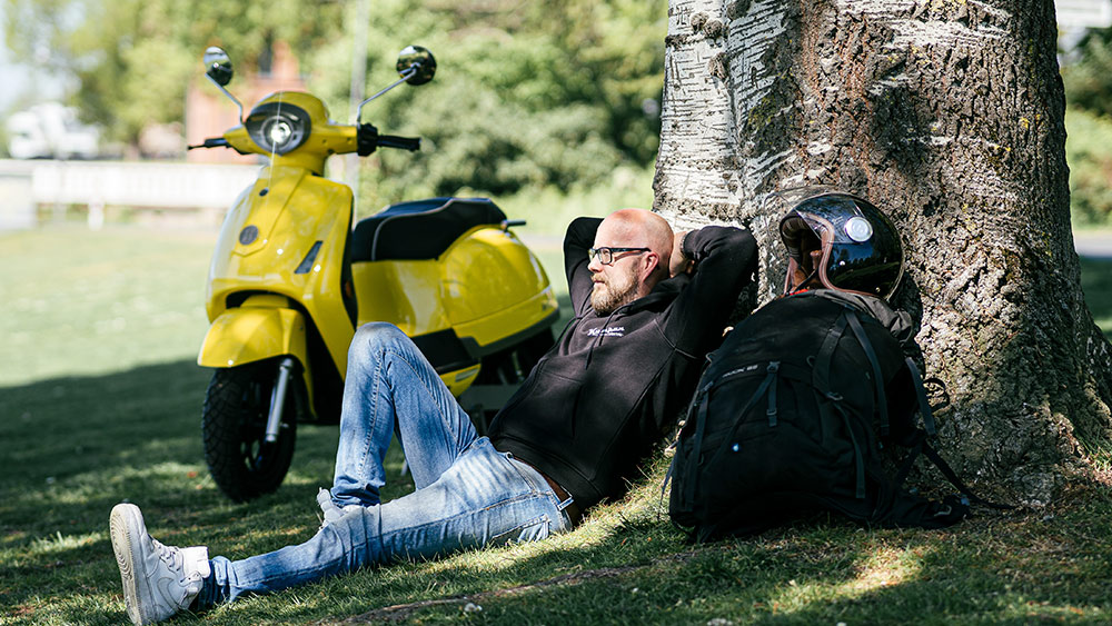 Man sitter på gräsmatta lutad mot ett träd med en gul moped bredvid sig