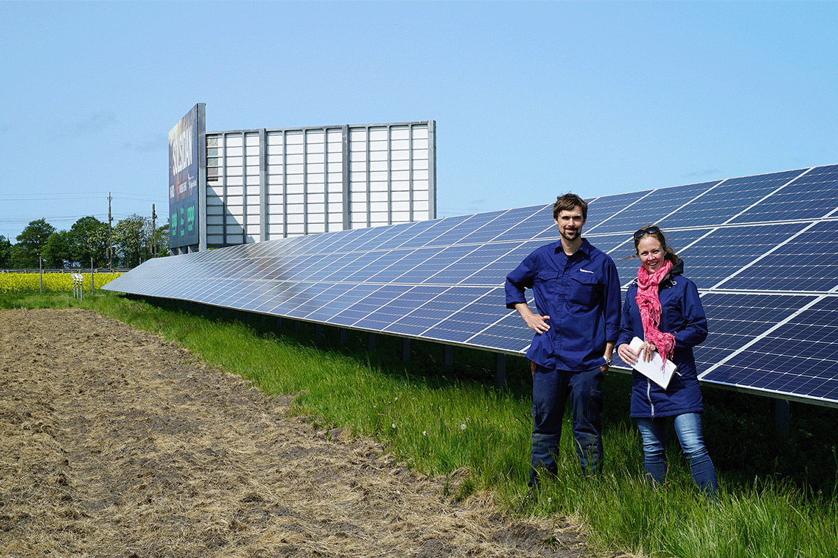 två personer poserar inför kameran framför stora solcellspaneler i en solcellspark