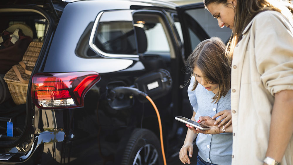mor o dotter står vid bilen och laddar den samtidigt som flickan tittar i mammans mobil