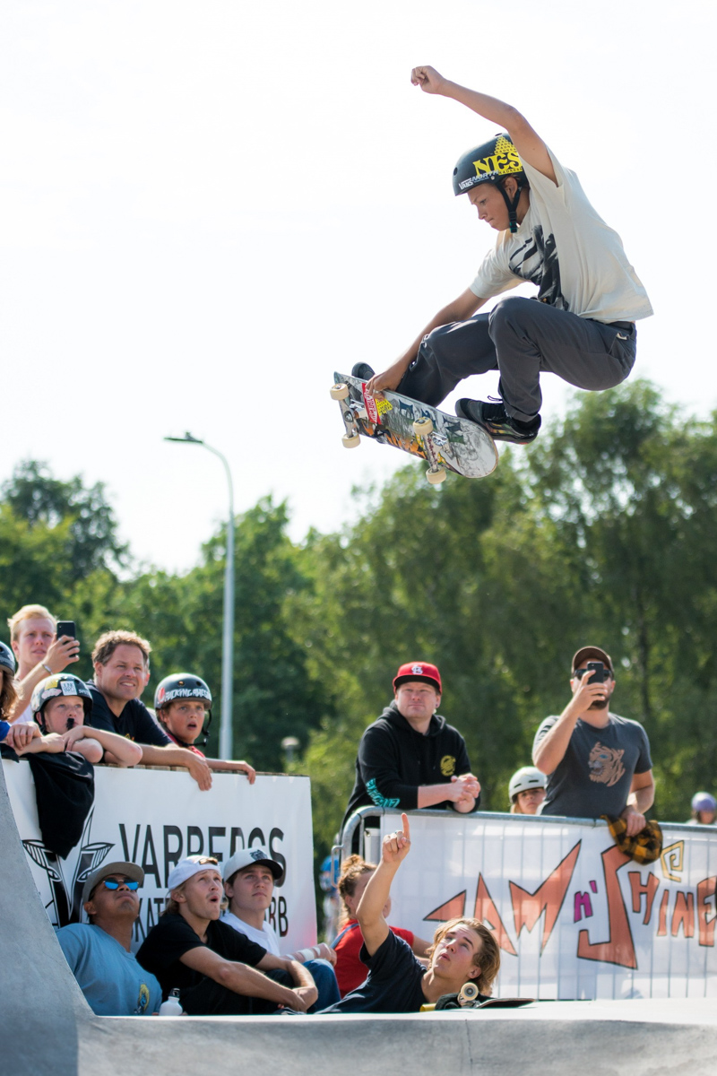 En ungkille hoppar högt med sin skateboard och flera ungdomar sitter nedanför och pekar glatt på honom